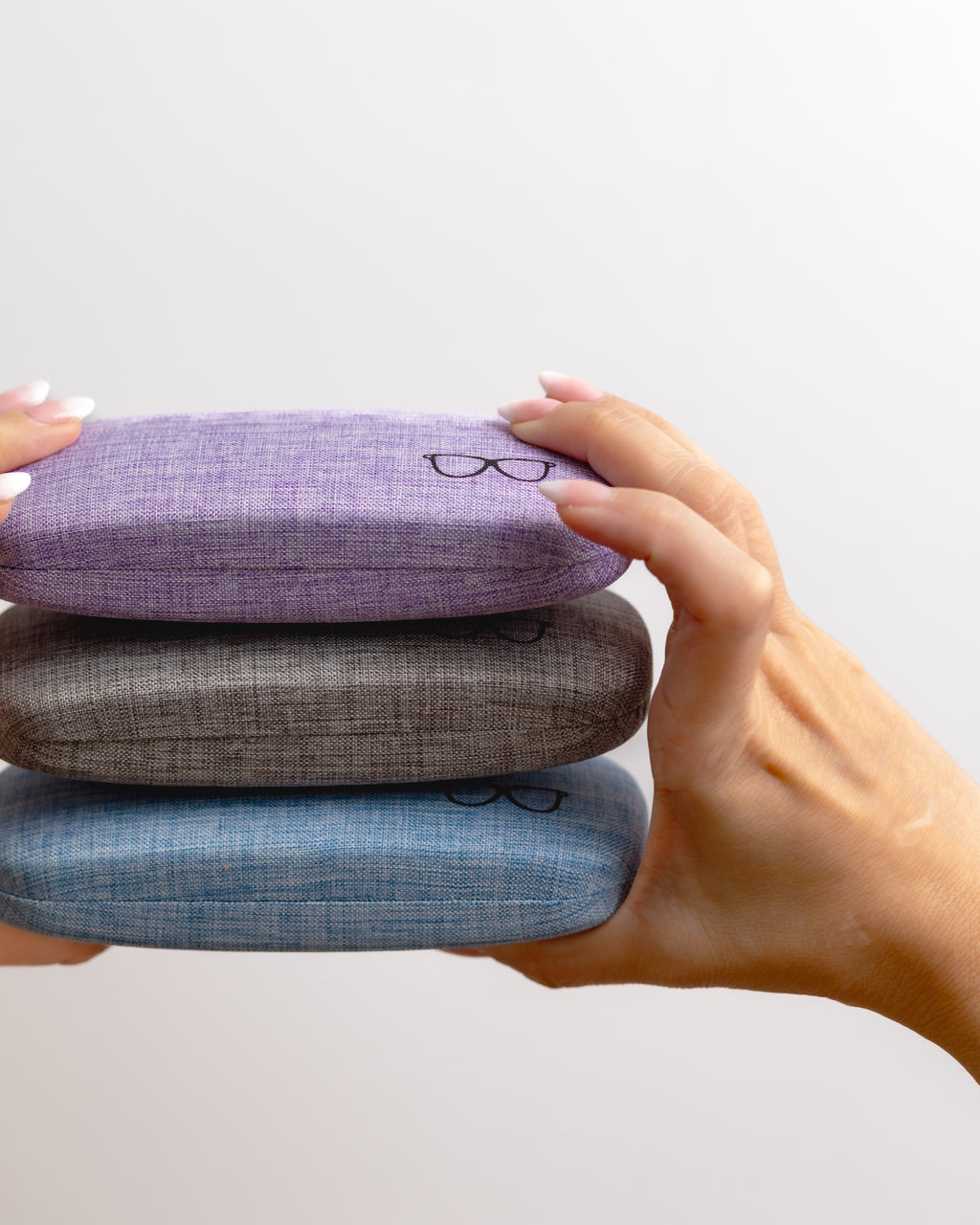 Three stacked eyeglass cases in purple, gray, and blue held by a hand against a light gray background.