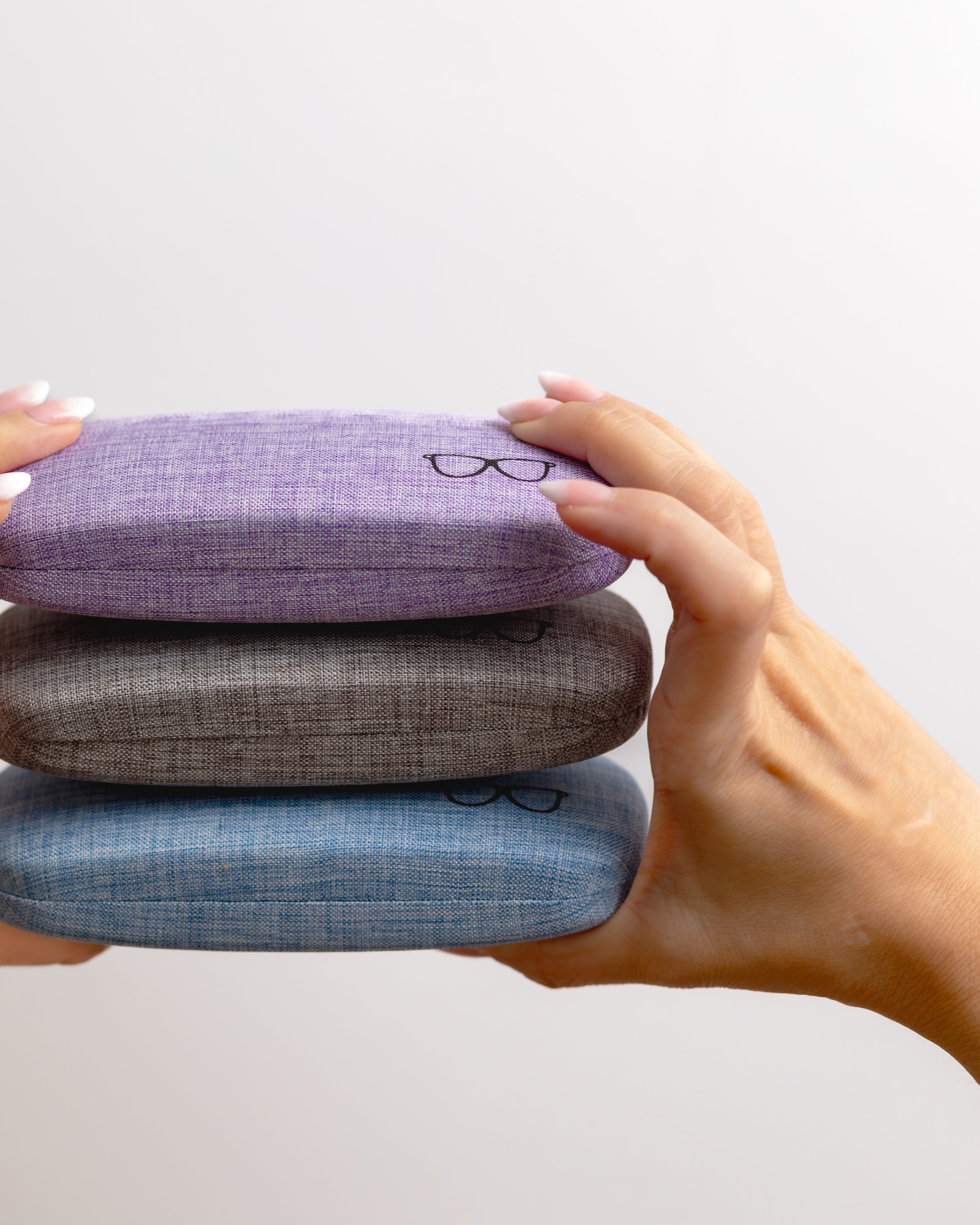 Three stacked eyeglass cases in purple, gray, and blue held by a hand against a light gray background.